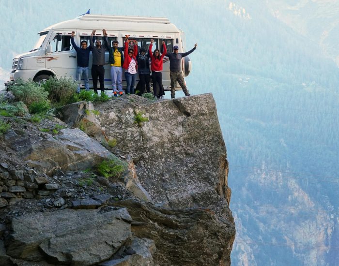 A group of friends celebrating on a cliff edge with a van in the background during a mountain adventure.